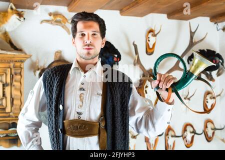 Giovane cacciatore con una tromba davanti a una parete con diversi corna, palchi e trofei in una baita alpina Foto Stock