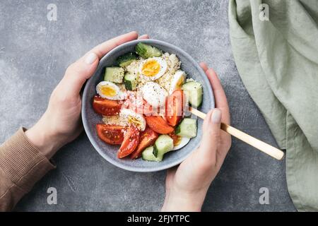 Le mani femminili tengono una ciotola di insalata di cous vegan con cetriolo, pomodoro di ciliegia e uova di quaglia su un tavolo di pietra. Foto Stock
