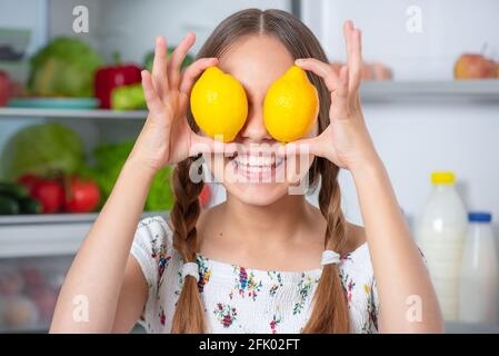 Sorridendo bella ragazza teen giovane che tiene i limoni gialli sopra gli occhi mentre si sta in piedi vicino al frigorifero aperto in cucina a casa Foto Stock