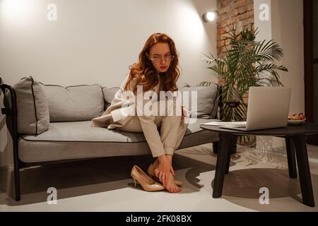 Giovane donna d'affari alla moda in un vestito da lavoro è stanca. Ha avuto una giornata ruvida, ha tolto i tacchi scomodi e le ha massaggiato le gambe. Foto Stock