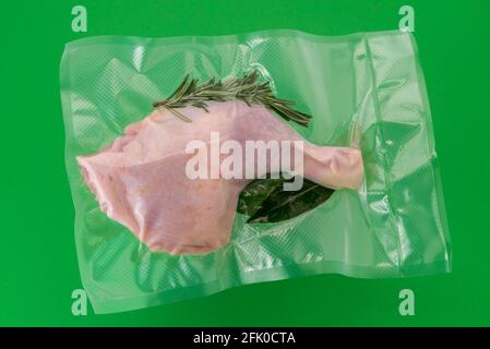 Cosce di pollo confezionate sottovuoto sigillate per cottura sous vide con rosmarino, foglia di alloro e salvia, isolate su sfondo verde Foto Stock