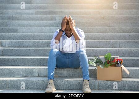 Crisi economica e bancarotta. Uomo nero disperato con oggetti personali seduti sulle scale dopo aver perso il lavoro, all'esterno Foto Stock