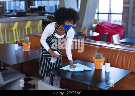 Cameriera donna afroamericana che indossa la maschera facciale, tavoli disinfettanti nel bar Foto Stock