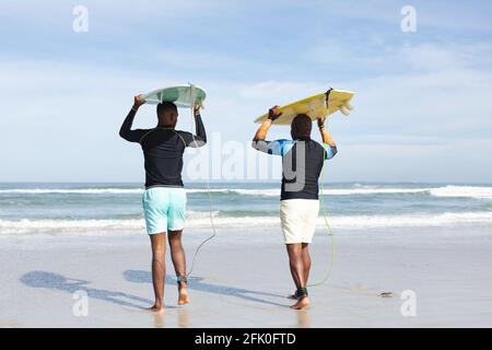 Vista posteriore del padre e del figlio afro-americani che trasportano tavole da surf sulla loro testa alla spiaggia Foto Stock