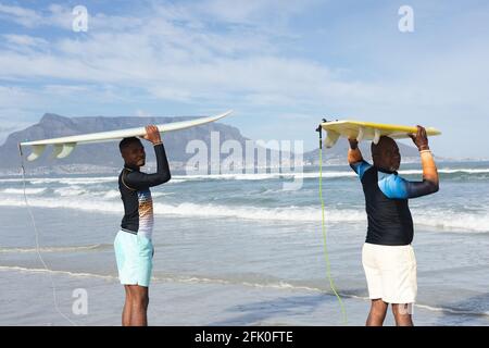Ritratto di padre e figlio afro-americano che porta tavole da surf la testa in spiaggia Foto Stock