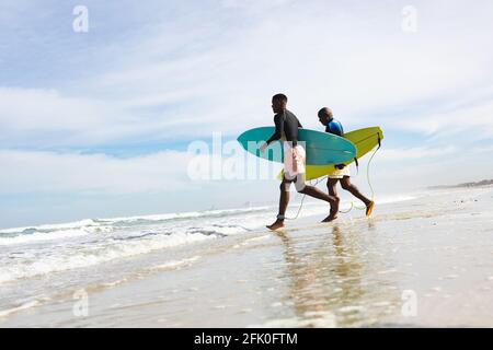Padre e figlio afro-americano con tavole da surf che corrono verso il onde in spiaggia Foto Stock