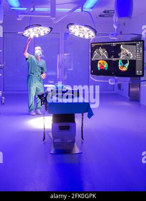 Essen, Germania. 27 Apr 2021. Un'immagine tomografica computerizzata di un cranio facciale può essere vista nella sala operatoria ENT dell'ospedale universitario, mentre la luce blu illumina la stanza e un uomo regola la lampada di funzionamento. Con l'aiuto della luce blu, i vasi sanguigni possono essere visualizzati meglio intraoperatoriamente. La nuova costruzione dell'ala operativa del teatro per le operazioni occhio e ENT costava quasi 70 milioni di euro. Secondo l'Ospedale Universitario, questi sono attualmente i teatri operativi più moderni in Germania e in Europa. Credit: Jonas Güttler/dpa/Alamy Live News Foto Stock