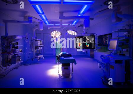 Essen, Germania. 27 Apr 2021. Un'immagine tomografica computerizzata di un cranio facciale può essere vista nella sala operatoria ENT dell'ospedale universitario, mentre la luce blu illumina la stanza e un uomo regola la lampada di funzionamento. Con l'aiuto della luce blu, i vasi sanguigni possono essere visualizzati meglio intraoperatoriamente. La nuova costruzione dell'ala operativa del teatro per le operazioni occhio e ENT costava quasi 70 milioni di euro. Secondo l'Ospedale Universitario, questi sono attualmente i teatri operativi più moderni in Germania e in Europa. Credit: Jonas Güttler/dpa/Alamy Live News Foto Stock