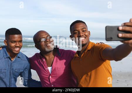 Il padre afroamericano e i suoi due figli che prendono un selfie dallo smartphone in spiaggia Foto Stock
