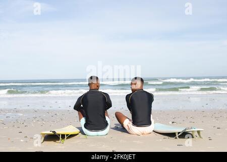 Vista posteriore dei fratelli afroamericani con tavole da surf sedute a. la spiaggia Foto Stock