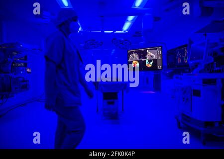 Essen, Germania. 27 Apr 2021. Un'immagine tomografica computerizzata di un cranio facciale è visibile nella sala operatoria ENT dell'ospedale universitario, mentre la luce blu illumina la stanza e un assistente chirurgico cammina attraverso la stanza. Con l'aiuto della luce blu, i vasi sanguigni possono essere visualizzati meglio intraoperatoriamente. La nuova costruzione dell'ala operativa del teatro per le operazioni occhio e ENT costava quasi 70 milioni di euro. Secondo l'Ospedale Universitario, questi sono attualmente i teatri operativi più moderni in Germania e in Europa. Credit: Jonas Güttler/dpa/Alamy Live News Foto Stock