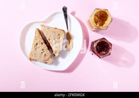 Piatto bianco a forma di cuore con panini con burro di arachidi e gelatina di fragole su sfondo rosa. Vista dall'alto Foto Stock