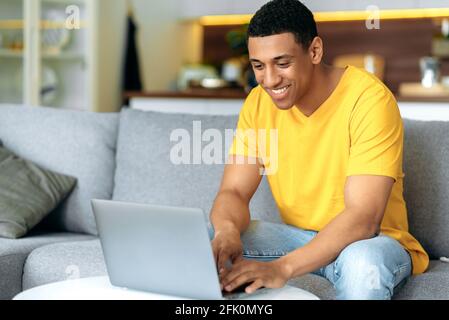 Giovane gioioso misto razza uomo moderno, freelance, seduto in soggiorno sul divano, utilizza un computer portatile, per lavorare a distanza, navigare in internet o chattare con gli amici su social network, sorridente amichevole Foto Stock