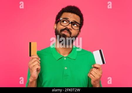 uomo afro-americano confuso in bicchieri che tengono le carte di credito isolate su rosa Foto Stock