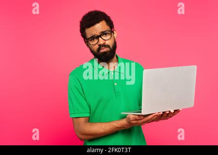 uomo afro-americano dispiaciuto in occhiali e polo verde camicia tenere il computer portatile isolato in rosa Foto Stock