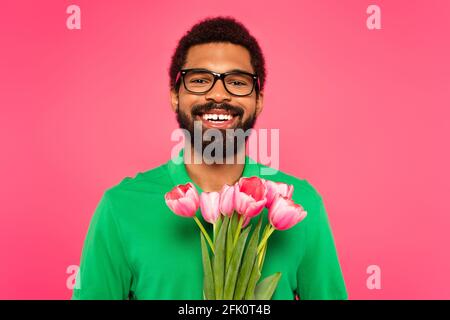 allegro uomo afroamericano in occhiali e polo verde camicia tenere tulipani isolati su rosa Foto Stock