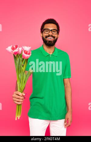 felice uomo afroamericano in occhiali e polo verde camicia tenere tulipani isolati su rosa Foto Stock