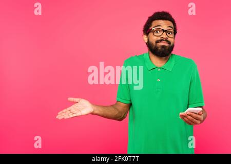 uomo afro-americano perplesso in una t-shirt verde che tiene isolato lo smartphone su rosa Foto Stock
