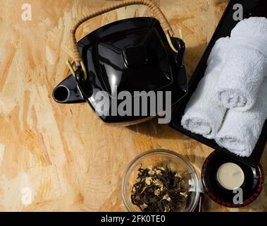 Una teiera nera con manico in vimini in stile orientale e rotoli di asciugamani bianchi in spugna su un piatto rettangolare nero. Tè flower sciolto in vetro. Foto Stock