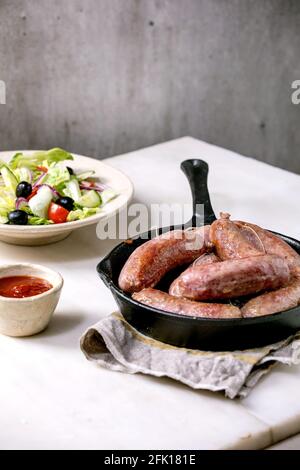 Salsiccia di salumi italiani alla griglia in padella di ghisa servita con salsa di pomodoro e piatto di insalata di verdure fresche su tavolo di marmo bianco. Cena bilanciata Foto Stock