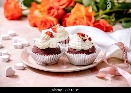 Cupcakes di velluto rosso fatto in casa con panna montata su piastra di ceramica rosa, tovagliolo bianco con nastro, fiori di rose, cuori di legno su fondo di tessitura rosa Foto Stock