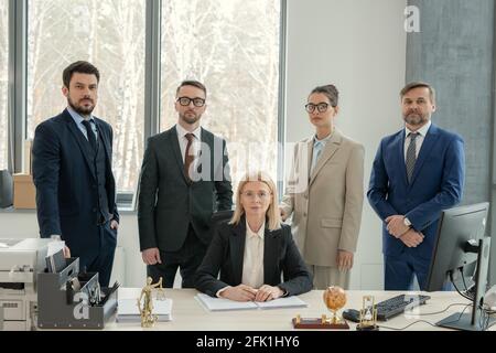 Serio cuoco femmina maturo di studio legale circondato da avvocati sedersi con documenti alla scrivania in un ufficio moderno Foto Stock