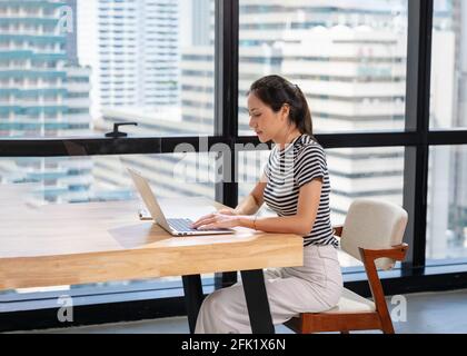 Bella donna d'affari asiatica in stoffa casual che lavora con la digitazione su computer portatile alla scrivania in un moderno ufficio vicino alla finestra Foto Stock