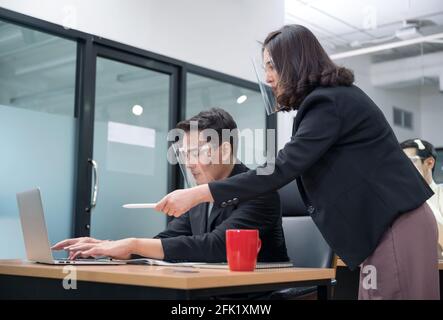 Bossy manager femmina ordinare e puntare al portatile di ufficio lavoratore maschile che indossa la visiera di protezione in un nuovo ufficio normale Foto Stock