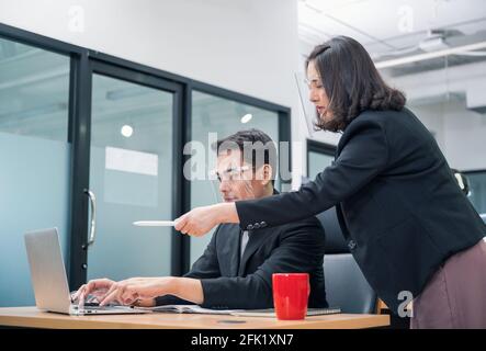 Bossy manager femmina ordinare e puntare al portatile di ufficio lavoratore maschile che indossa la visiera di protezione in un nuovo ufficio normale Foto Stock
