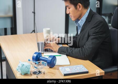 Asian marketing manager uomo che lavora con laptop e prodotto piggy banca, dumbbell, mini carrello di shopping su scrivania in legno in ufficio Foto Stock