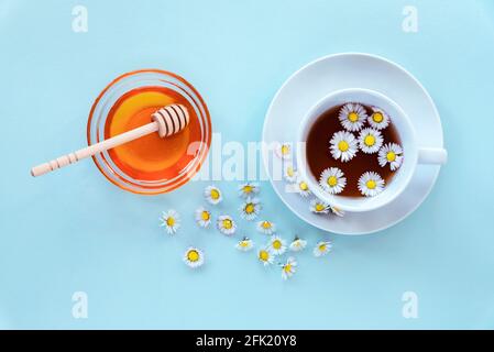 Una tazza di tè con camomilla e miele in una ciotola di vetro con cucchiaia di legno su sfondo blu. Vista dall'alto, disposizione piatta. Foto Stock