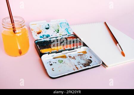 Taccuino bianco di colore dell'acqua, l'arte fornisce i pennelli, le vernici, i acquerelli e la tazza di acqua. Studio d'arte. Lezioni di disegno. Foto Stock