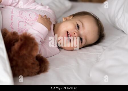 Autentico ritratto cute caucasica bambino bambino bambino chubby ragazza o. ragazzo in rosa sonnolento al risveglio con orsacchiotto che guarda a camera in letto bianco Foto Stock