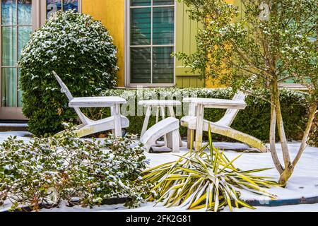 Due sedie Adirondack bianche con un tavolo tra loro la neve di fronte a una casa colorata con verde persiane e arbusti intorno a loro Foto Stock