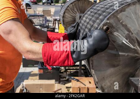 Il lavoratore arresta il torrefattore di chili del barile caldo dal girando e prepara per aprirla indossando i vertici del forno per impieghi pesanti all'aperto tratteggio di un evento di arrostimento del peperoncino Foto Stock