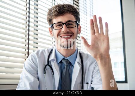 Sorridente giovane medico maschio che agitava la mano alla macchina fotografica salutando il paziente online tramite videochiamate a distanza dall'ufficio Foto Stock