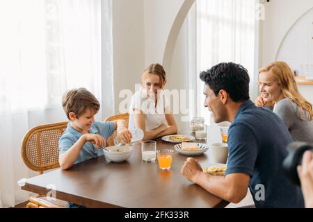 Bel giovane padre con bella madre e figlia teen guardando il bambino piccolo che versa il latte dal biberon nel recipiente di cereali durante la prima colazione a casa Foto Stock