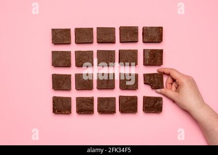Vista dall'alto con la mano di una donna che afferra un pezzo di torta al cioccolato fondente. Disposizione piatta con barrette di fondente di cioccolato disposte simmetricamente su uno sfondo rosa Foto Stock