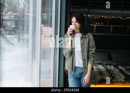 Pensieroso bella donna di mezza età in piedi in una camera da letto accanto a una finestra, bere caffè, non guardando da nessuna parte. Indossa abiti casual. Foto Stock