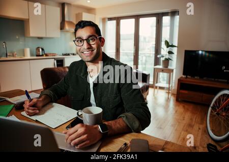 Uomo d'affari sorridente che scrive sul blocco note godendo il caffè caldo seduto a. ufficio domestico Foto Stock