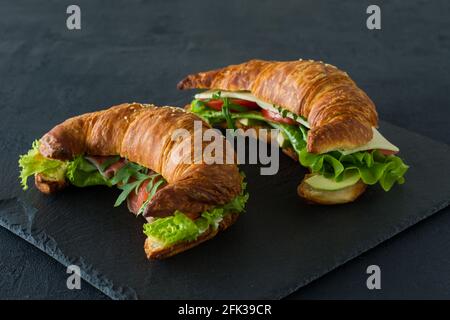 Panini con croissant e salmone salato su una scrivania, serviti con foglie di insalata fresca, rucola e verdure su sfondo nero. Foto Stock
