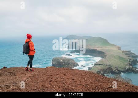 Donna vestita con una felpa con cappuccio arancione e cappuccio rosso con zaino che gode di vista sull'oceano Atlantico sulla penisola di Ponta de Sao Lourenco Foto Stock