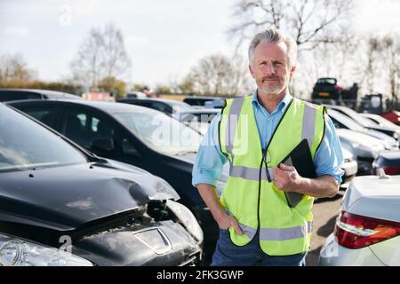 Ritratto del regolatore di perdita maschio maturo che indossa un gilet di sicurezza ad alta visibilità in piedi in composto per auto danneggiate compilare il rapporto di reclamo di assicurazione nella scheda digitale Foto Stock