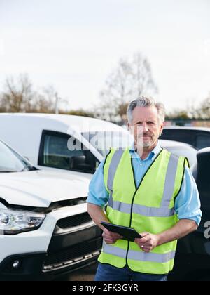 Ritratto del regolatore di perdita maschio maturo che indossa un gilet di sicurezza ad alta visibilità in piedi in composto per auto danneggiate compilare il rapporto di reclamo di assicurazione nella scheda digitale Foto Stock