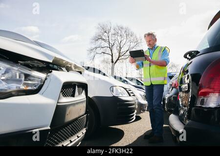 Regolatore di perdita maschio maturo che indossa gilet di sicurezza ad alta visibilità in piedi composto per auto danneggiate che scattano foto per rapporto di reclamo di assicurazione su tablet digitale Foto Stock