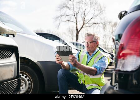 Regolatore di perdita maschio maturo che indossa gilet di sicurezza ad alta visibilità in piedi composto per auto danneggiate che scattano foto per rapporto di reclamo di assicurazione su tablet digitale Foto Stock