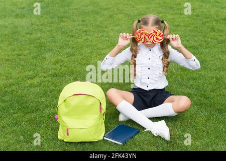 Bambina in uniforme scolastica divertirsi coprendo gli occhi con lollipop su erba verde, dente dolce Foto Stock