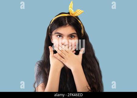 Scioccata ragazza teen indiana che copre la bocca con le mani e guardando la macchina fotografica, mantenendo il silenzio su sfondo blu Foto Stock