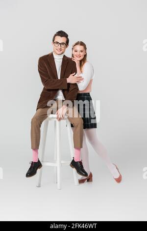 donna felice in gonna di plaid appoggiata sulla spalla dell'uomo seduta su sgabello alto su sfondo grigio Foto Stock