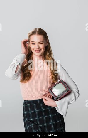 donna flirty con tv vintage che fissa i capelli mentre winking a. telecamera isolata in grigio Foto Stock